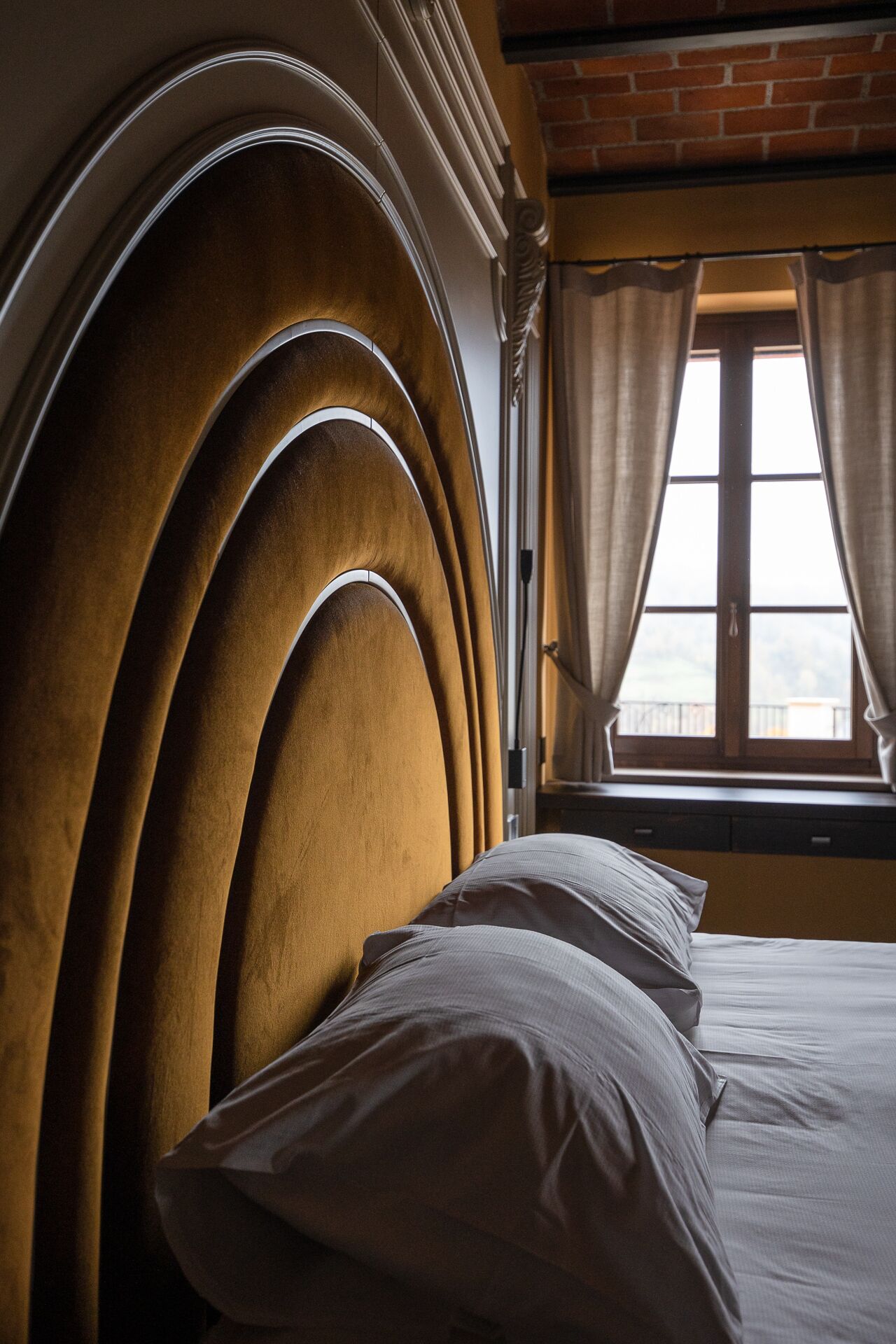A bed with a curved headboard, a window with curtains, and a brick wall in the background.
