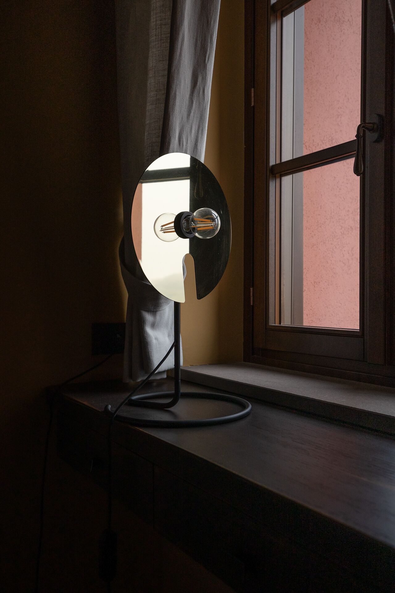 A desk lamp with a reflective surface, positioned on a desk near a window with a view of a building.