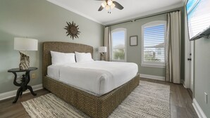 Une chambre avec un grand lit, un ventilateur de plafond, un téléviseur fixé au mur et une fenêtre avec des rideaux.