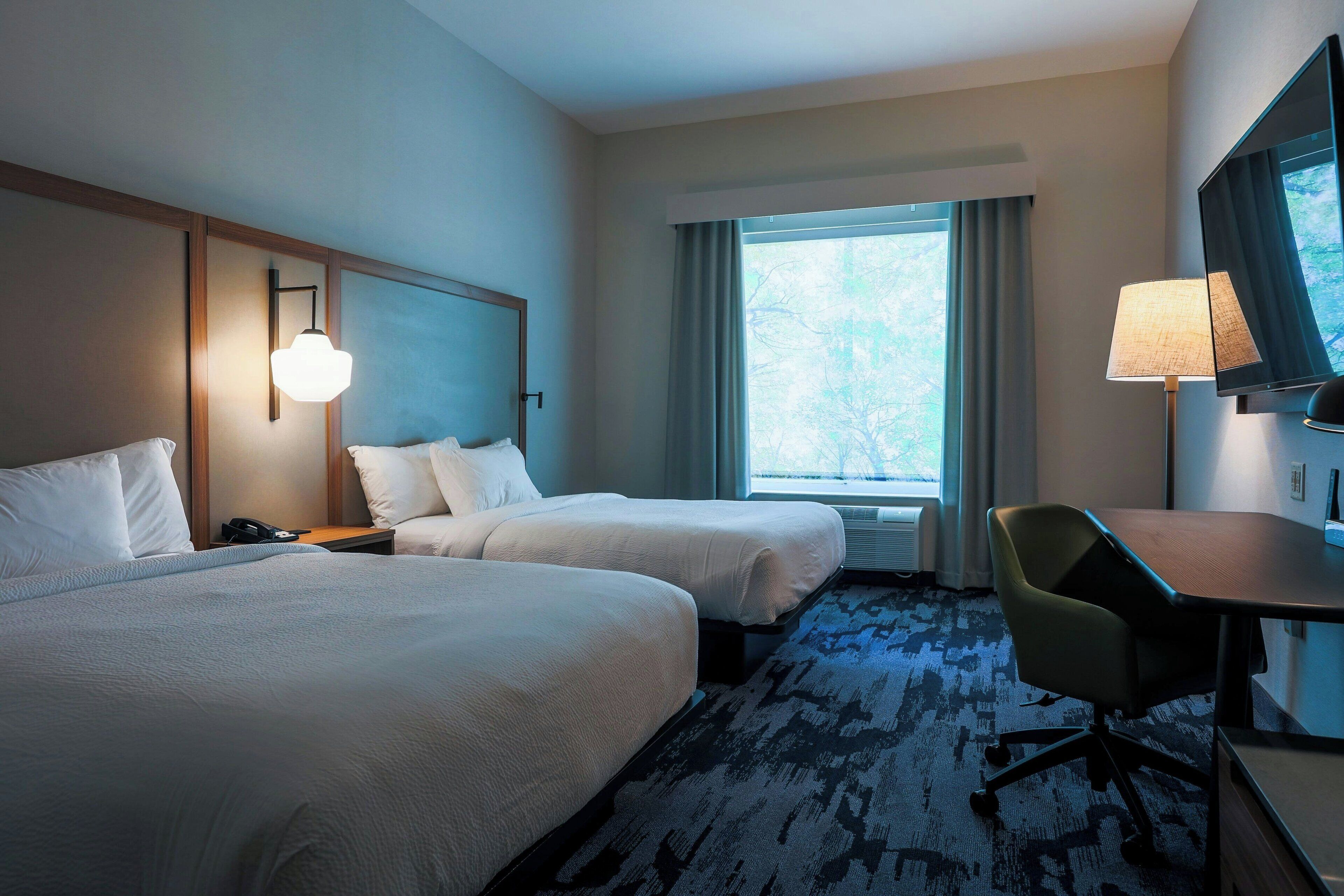 A hotel room with two beds, a desk, a chair, a TV, and a window with curtains.