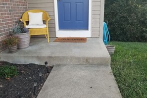 A concrete front porch with a blue door, a yellow wicker chair with a white cushion, and potted plants.