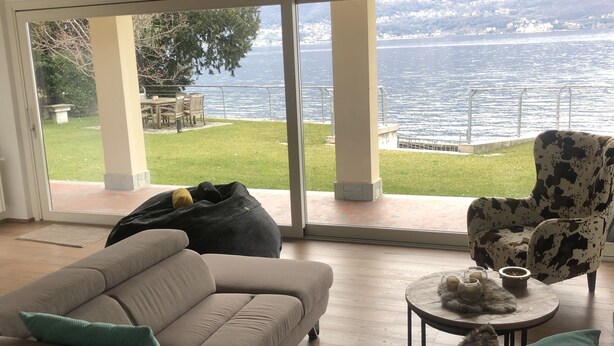 A modern living room with a large sofa, a patterned armchair, and a small round table with candles. A dog is visible in the foreground. The room has large sliding glass doors leading to a patio with a view of a body of water and greenery. - Remarkable 4-bed Villa in Bellano (Bellano)