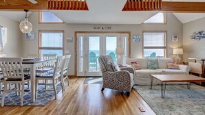 A spacious living area with a dining table, chairs, a patterned armchair, a sofa, and a coffee table. There are ceiling fans, a view of the ocean through large windows, and a sign reading "SURF CITY". - Oceanfront Getaway In Surf City (Surf City)