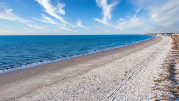 A wide sandy beach with gentle waves, a clear sky, and a distant shoreline with buildings. - One of kind 3 bedroom Villa with Ocean/Lagoon Views. (Hilton Head Island)