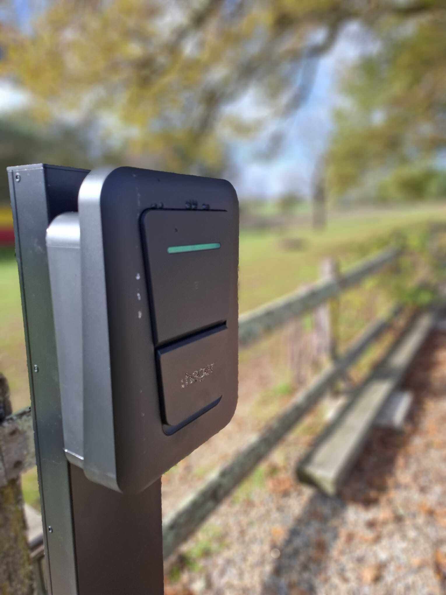 Um dispositivo eletrônico preto e prateado fixado em um poste de metal, provavelmente um parquímetro ou máquina de bilhetes, com uma luz indicadora verde.