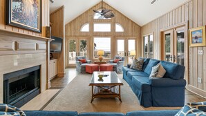 A spacious living room with a fireplace, a blue sofa, and a large window offering a view of the outdoors. - Beachside Treehouse, spectacular ocean view on popular Surfsong Rd (Kiawah Island)