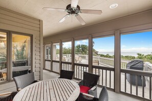 A covered outdoor deck with a round wooden table and wicker chairs, large windows, and a view of the ocean.