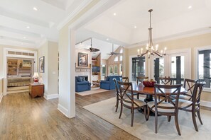 A spacious dining area with a wooden table and chairs, a chandelier, and a view of the kitchen and living room through large windows.