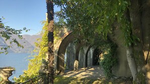 A pathway with stone arches leading to a body of water, surrounded by trees and grass. - Villa Giudici Luxury app on the Lake (Nesso)