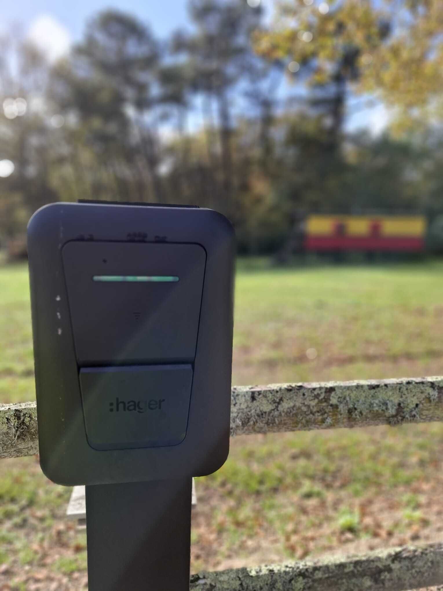Un appareil électronique noir, fixé sur un poteau, avec un voyant lumineux vert, se détache sur un arrière-plan flou d’un parc, où l’on aperçoit un objet rouge et jaune au loin.