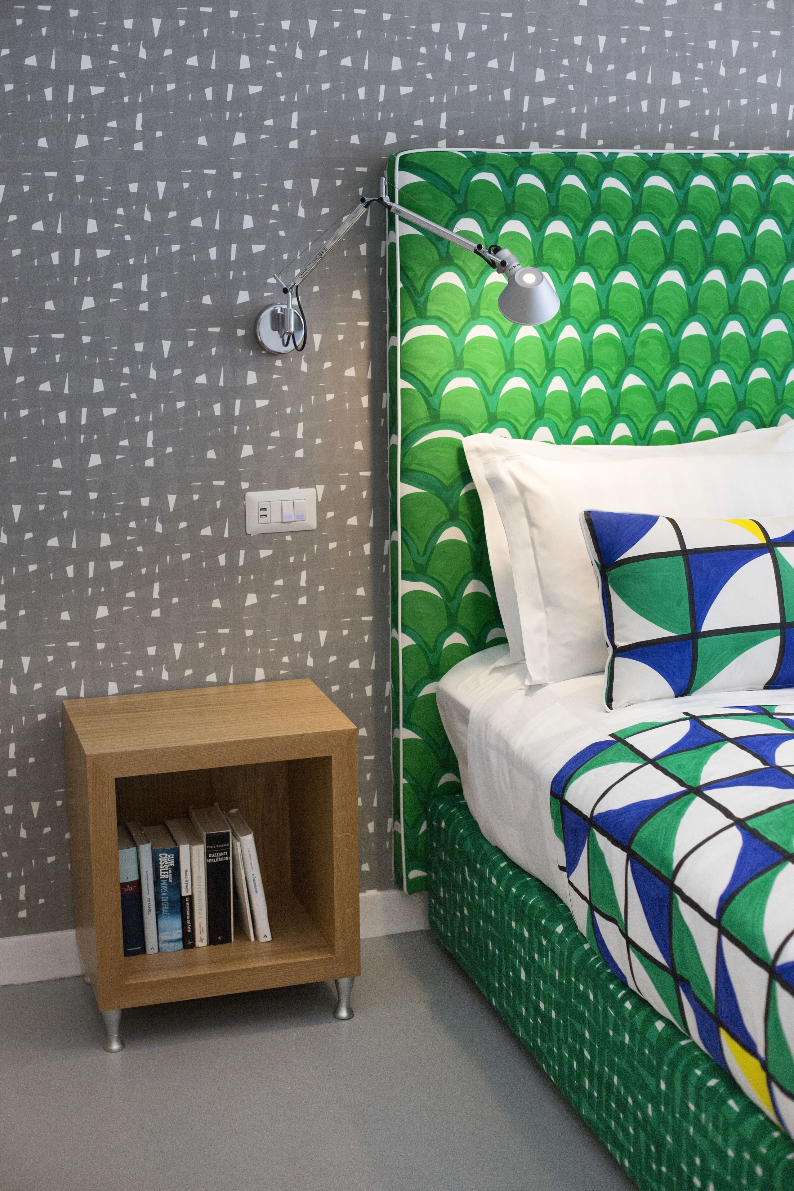 A bedroom with a green patterned headboard, a bedside table with books, and a lamp.
