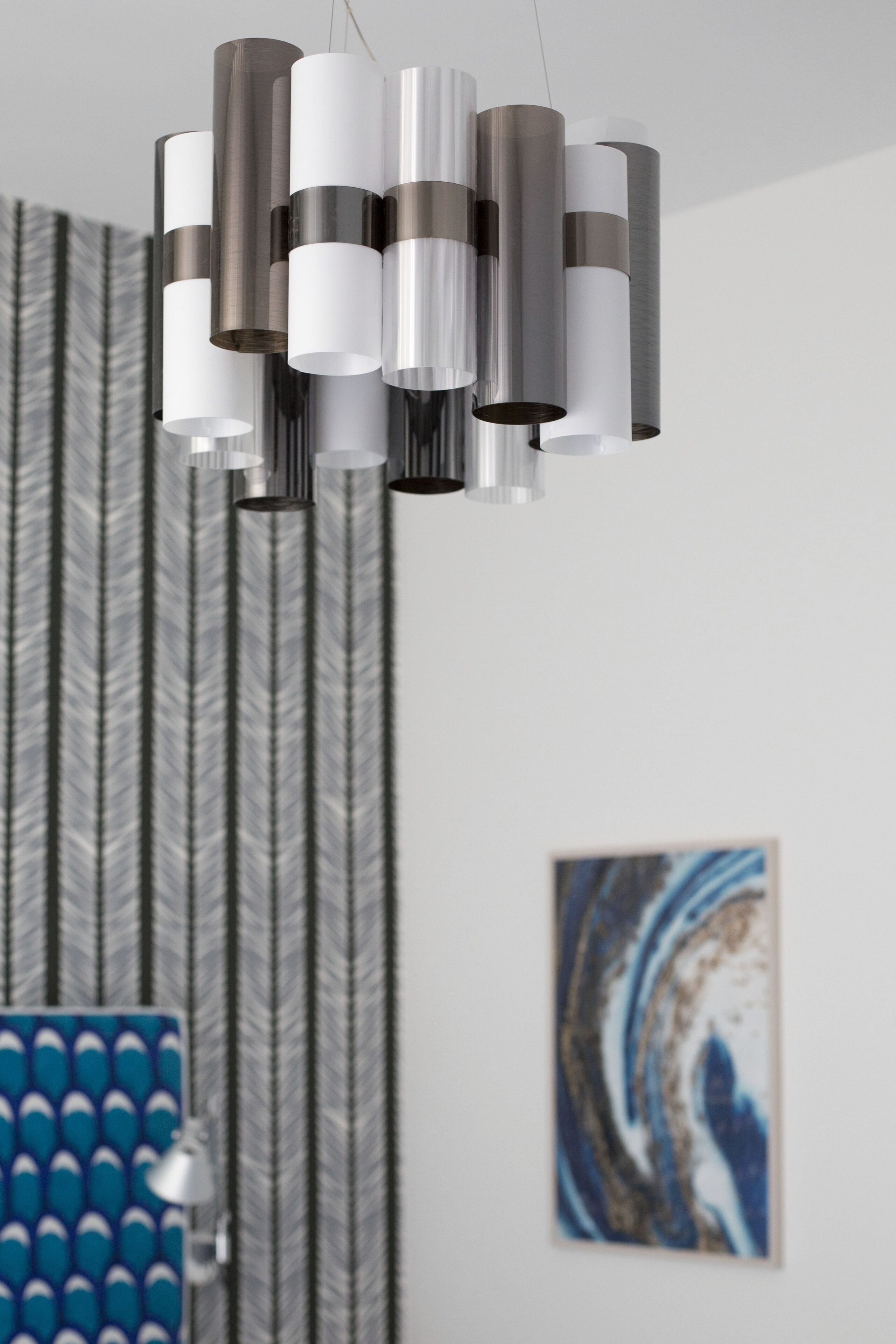 A modern chandelier with multiple cylindrical shades in a room with patterned wallpaper and a framed abstract painting.
