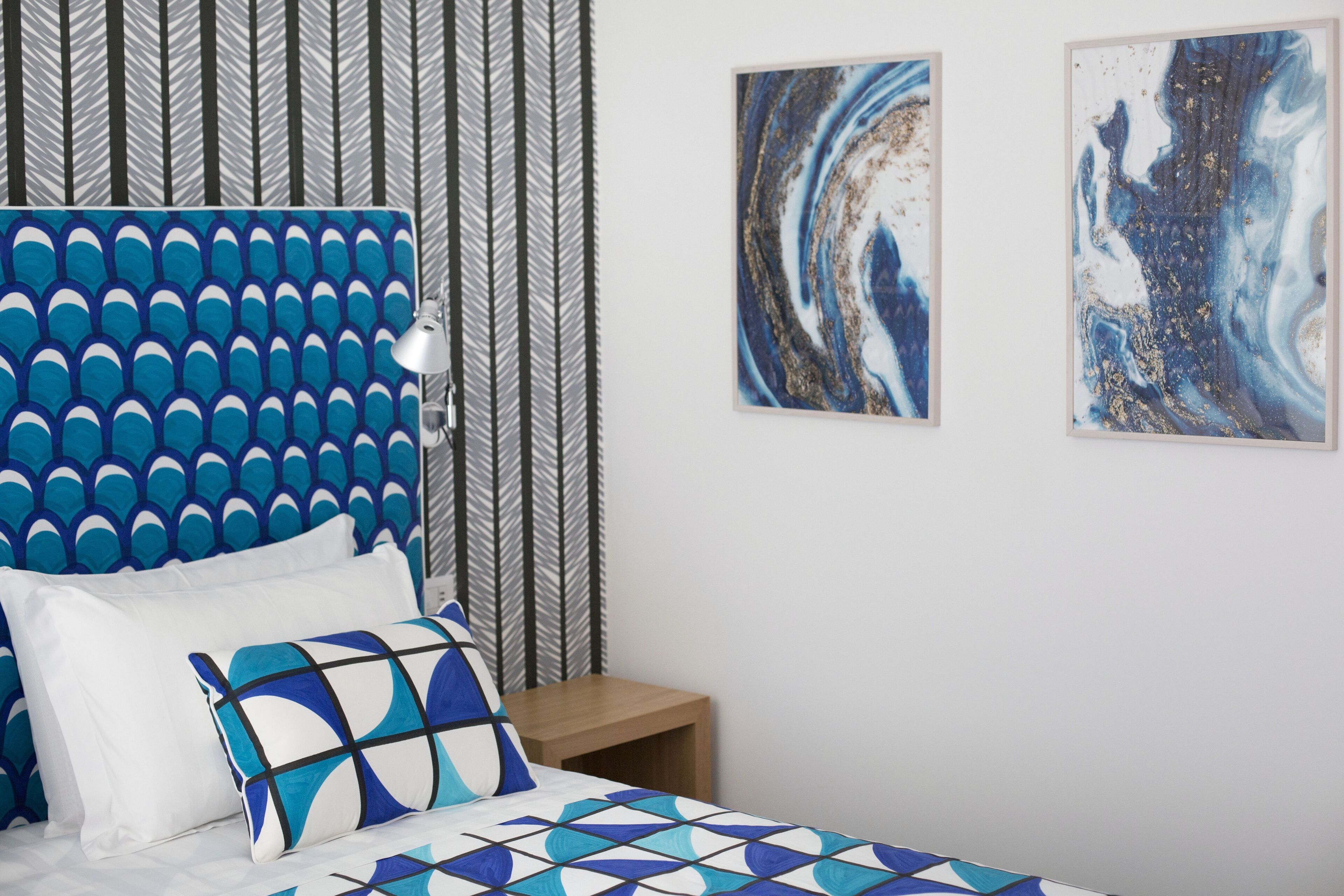 A bedroom with a patterned headboard, a bed with a geometric-patterned bedspread, two pillows, a bedside table, and two abstract wall art pieces.