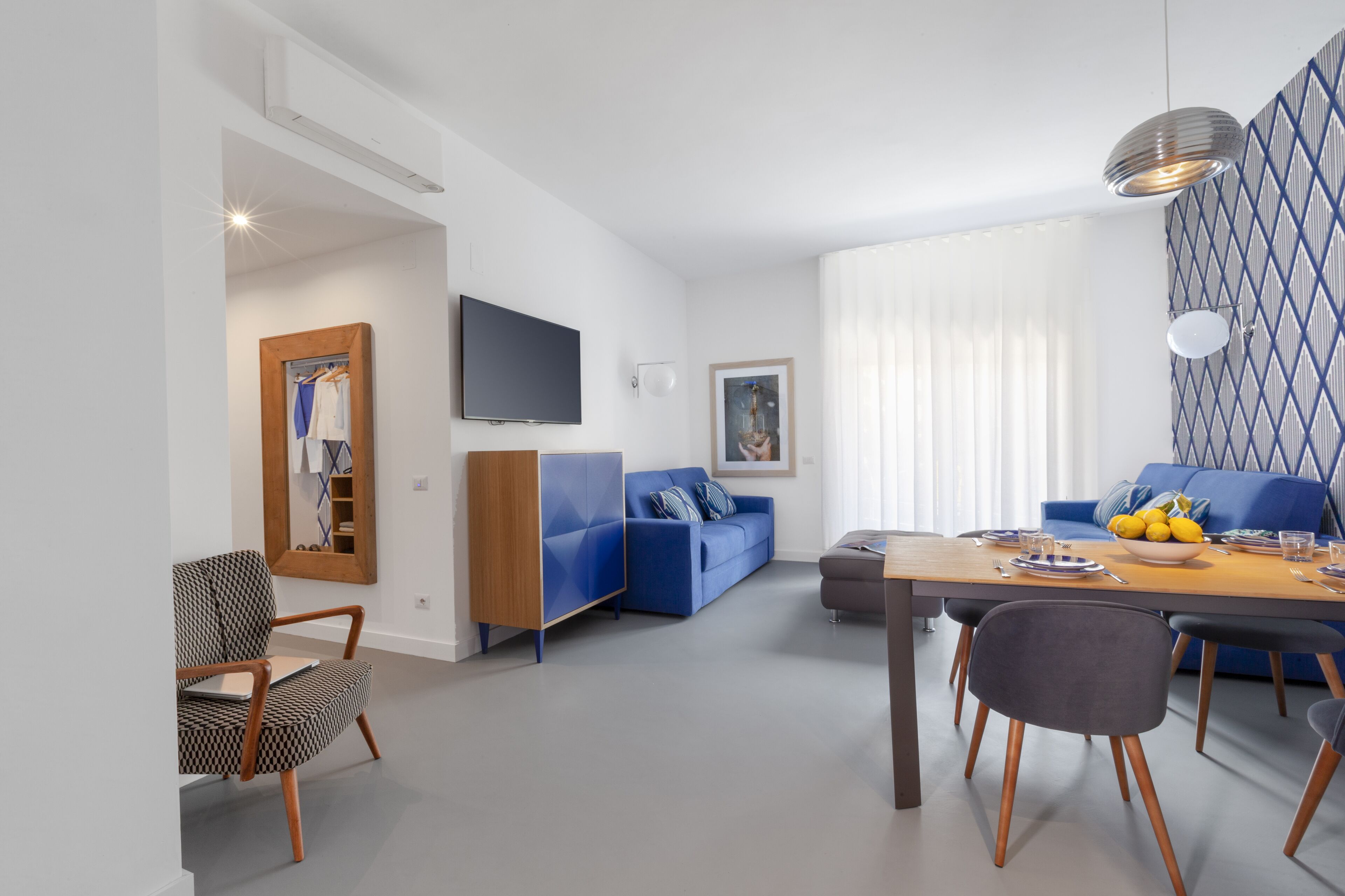 A modern living room with a blue sofa, a wooden dining table, and a TV mounted on the wall.