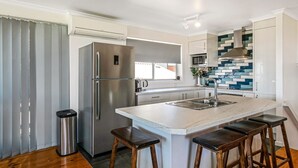A modern kitchen with a stainless steel refrigerator, a built-in oven, a microwave, and a sink with a tiled backsplash. There are four wooden stools around the island. - Goku's Shack - Rural and water views with a spa (CORONET BAY)