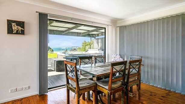 A dining area with a wooden table and chairs, a view of the sea, and a framed picture on the wall. - Goku's Shack - Rural and water views with a spa (CORONET BAY)