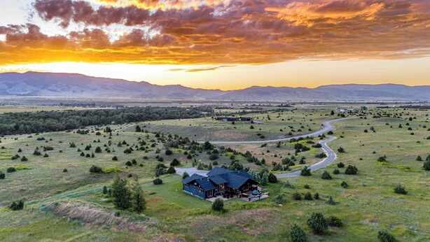 Une grande maison moderne dotée d’un vaste patio, entourée d’un paysage ouvert et étendu, avec une route sinueuse et des montagnes à l’horizon.