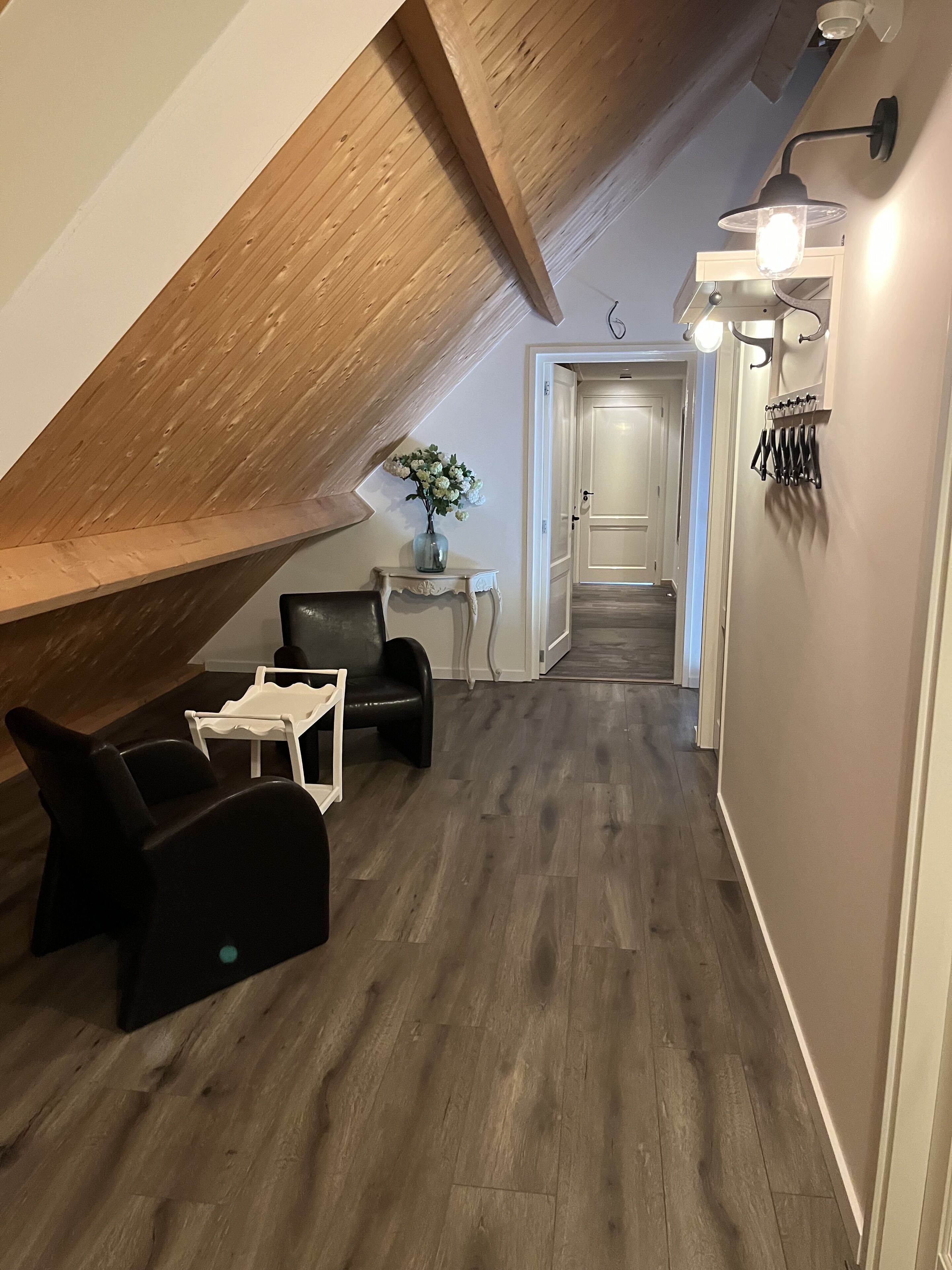 Een loftachtige kamer met een schuine plafond, een klein tafeltje met een bloemenvaas, een bank en een stoel.