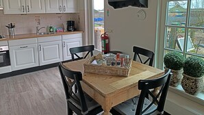 Une cuisine avec des meubles blancs, une table en bois avec des chaises, un panier rempli d’objets et une fenêtre ornée de plantes.