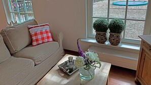 Un salon chaleureux avec un canapé beige, un coussin à carreaux rouges et blancs, une petite table sur laquelle est posé un vase de fleurs, et une fenêtre donnant sur un terrain de jeux et un trampoline.