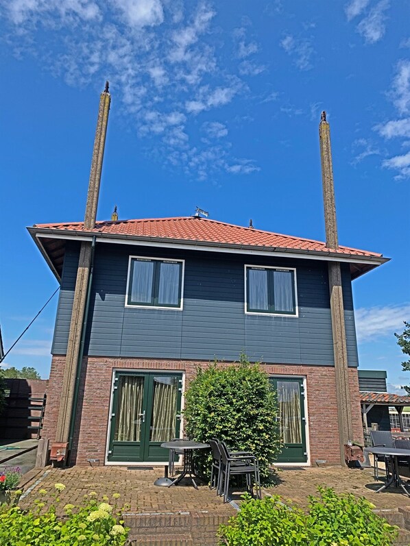 Un edificio de dos pisos con techo de tejas rojas, dos chimeneas y fachada de ladrillo. Cuenta con un pequeño patio con muebles de exterior y un arbusto verde.