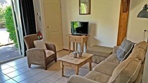 A living room with a sofa, a wicker chair, a small wooden table with two cups, a television on a stand, and a door leading outside. - Christinahoeve Oude Deel #1 (Boskoop)