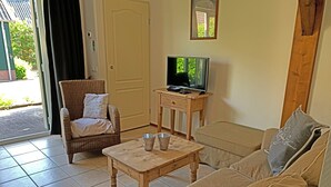 Ein Wohnzimmer mit einer Couch, einem Korbstuhl, einem kleinen Holztisch mit zwei Tassen, einem Fernseher auf einem Holzschrank und einer Tür mit Fenster.