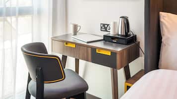 A hotel room with a desk, a chair, a bed, a coffee maker, and a wall-mounted power outlet.