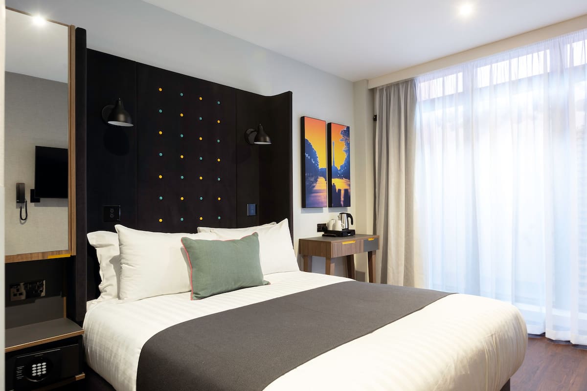 Une chambre d’hôtel avec un grand lit, une table de chevet avec une lampe, un tableau au mur et une fenêtre avec des rideaux.