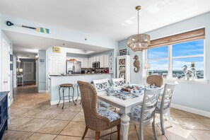 A spacious kitchen with white cabinets, a dining area with a white table and wicker chairs, and a view through large windows. - Deluxe Views-Short Walk 2 Beach-Resort Amenities (Miramar Beach)