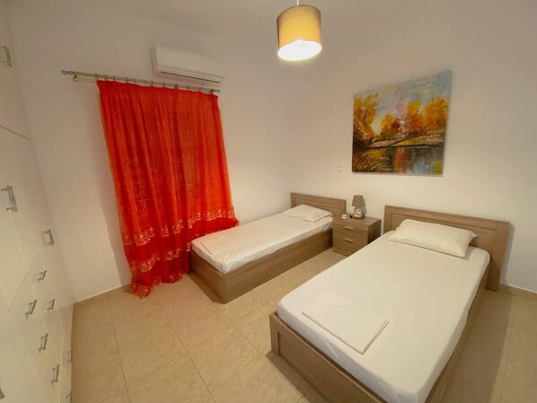 A hotel room with two beds, a nightstand, an air conditioning unit, a red curtain, and a painting on the wall.