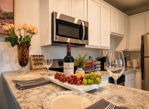 Une cuisine avec des meubles blancs, un four à micro-ondes en acier inoxydable, une bouteille de vin et un plateau de fruits.