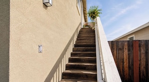 A narrow staircase leading to a building with a beige exterior wall, a wooden fence, and a potted plant with yellow flowers at the top. - Sonoma Wine Country Retreat | Near Top Wineries + Cozy Escape (Santa Rosa)
