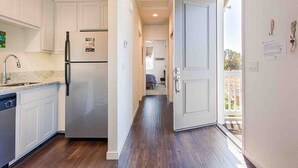 A narrow kitchen with white cabinets, a stainless steel refrigerator, and a dishwasher. A hallway leads to a bedroom with a bed and a chair. - Sonoma Wine Country Retreat | Near Top Wineries + Cozy Escape (Santa Rosa)