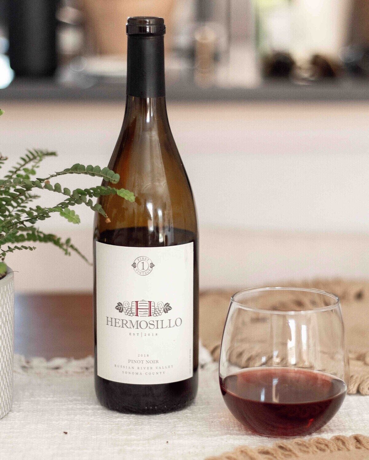 A bottle of Hermosillo Pinot Noir wine next to a glass filled with red wine, placed on a textured surface.