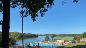 En swimmingpool med liggestole og parasoller, omgivet af et hegn, med udsigt til en sø og bakker i baggrunden.