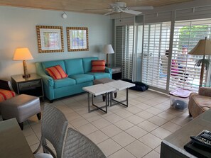 A room with a teal sofa, a small table, a chair, a ceiling fan, and a window with blinds. - Gulf Beachfront 2BR Condo with Golf Privileges  (Sanibel)