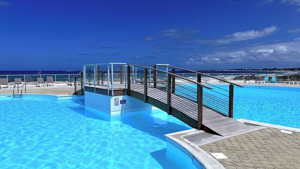 Uma piscina com um deque de madeira e cadeiras de descanso, sob um céu azul límpido.