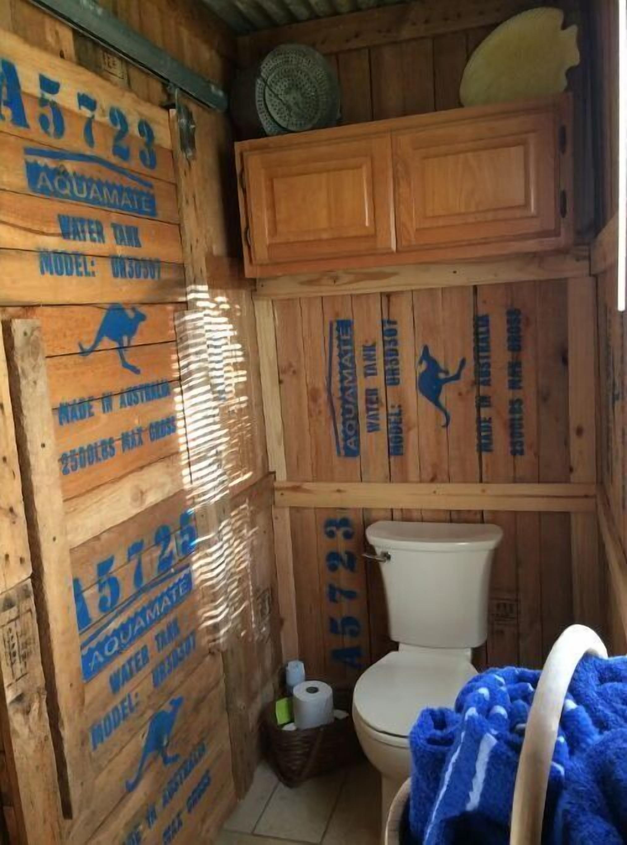 A rustic wooden bathroom with a toilet, a fan, and a basket of toiletries.