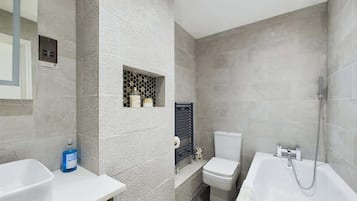 Un baño moderno con bañera, inodoro y lavamanos, que presenta una pared con textura y un pequeño estante con artículos de aseo.