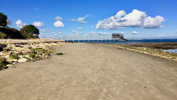 Una playa con un muelle que se adentra en el mar, un edificio sobre el muelle y un litoral rocoso.