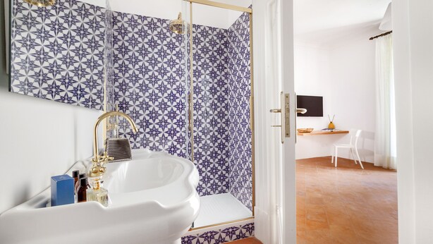 Um banheiro com pia branca, chuveiro com azulejos estampados em azul e uma cadeira branca no quarto vizinho.
