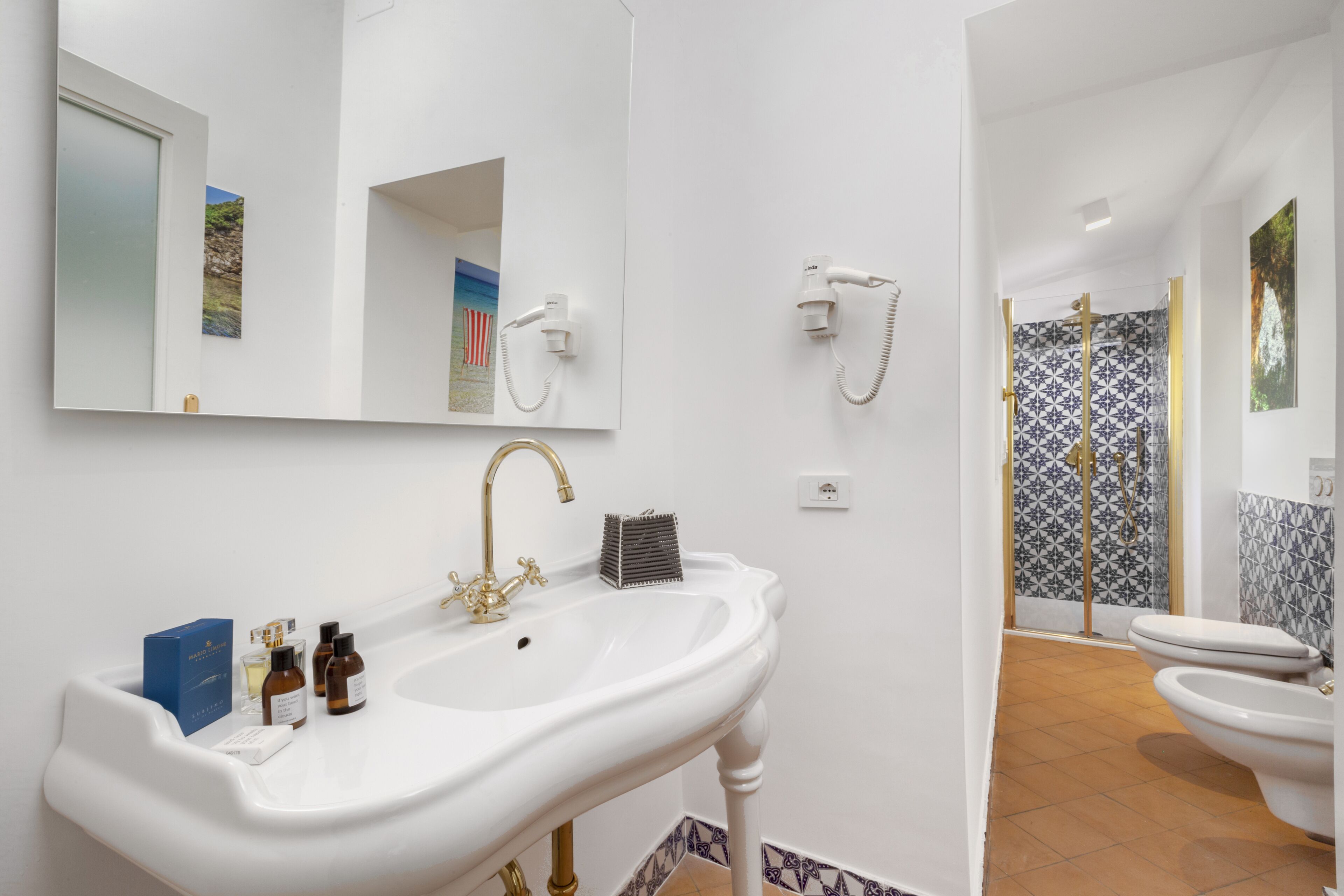 A bathroom with a white sink, gold faucet, and a mirror above it. There are two hair dryers mounted on the wall, and a shower with patterned tiles.