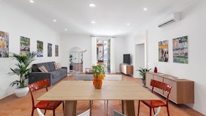 A spacious living area with a wooden dining table, red chairs, a grey sofa, and a TV on a wooden cabinet.