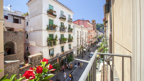 A narrow street with buildings on both sides, balconies with plants, and a clear blue sky.