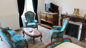 A room with two blue upholstered armchairs, a wooden coffee table, a television on a wooden cabinet, and a fireplace with decorative items. - JOSEPHINE BEDROOM  (Luxeuil-les-Bains)