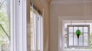 A room with a large window, a smaller window with a stained glass design, and a wooden chair.