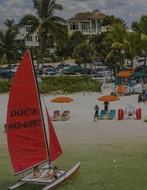 Un velero con vela roja en una playa con palmeras y un edificio al fondo.