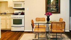 A modern kitchen with white cabinets, a stove, and a microwave. There is a dining area with wicker chairs and a vase of flowers. A painting hangs on the wall. - Montrose Place #4 Sum (Houston)
