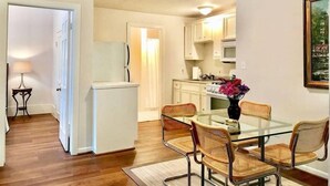 A compact kitchen with white cabinets, a refrigerator, and a dining area with a glass table and wicker chairs. - Montrose Place #4 Sum (Houston)
